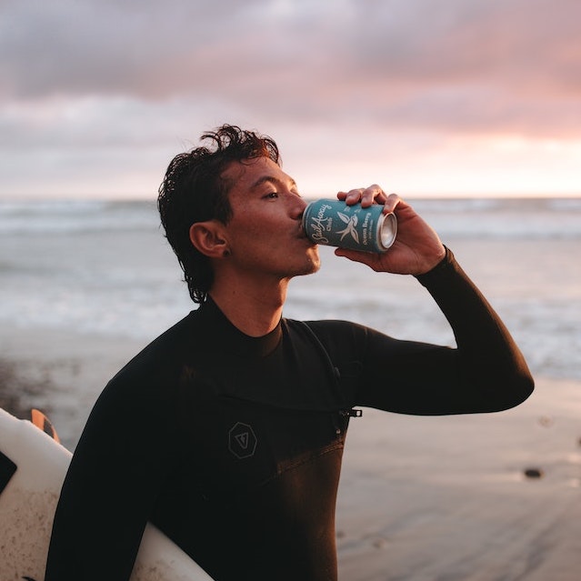 A man with a surf board drinking from a can